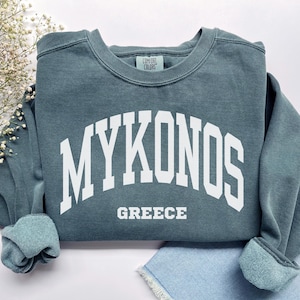 Puede incluir: Una sudadera de color verde azulado con la palabra "MYKONOS" en letras blancas grandes arqueadas sobre el pecho, con "GREECE" impresa debajo. La sudadera tiene cuello redondo y puños enrollados.
