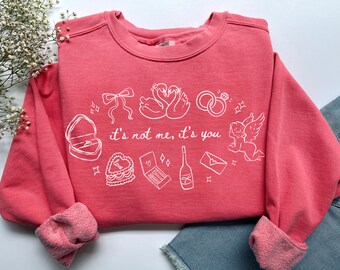 May include: A coral-colored sweatshirt with white graphic illustrations of a ring box, swans, rings, a cake, matches, a champagne bottle, an envelope, a bow, and an angel. The text "it's not me, it's you" is printed in white.