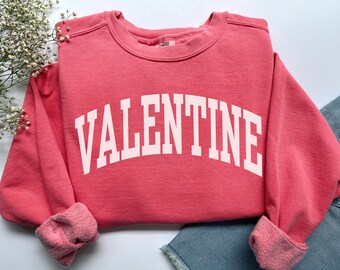May include: A coral pink crewneck sweatshirt featuring the word VALENTINE printed in large, arched, white block capital letters across the chest. The garment has long sleeves with ribbed cuffs and a soft, fleece-lined interior texture.