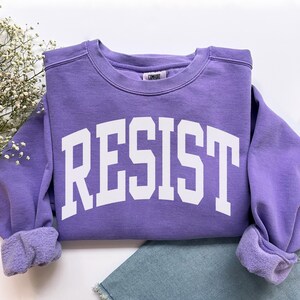 May include: A lavender crewneck sweatshirt with the word "RESIST" in bold white letters. The sweatshirt has a comfortable, casual style, with cuffed sleeves and a relaxed fit. The garment is paired with blue denim jeans.