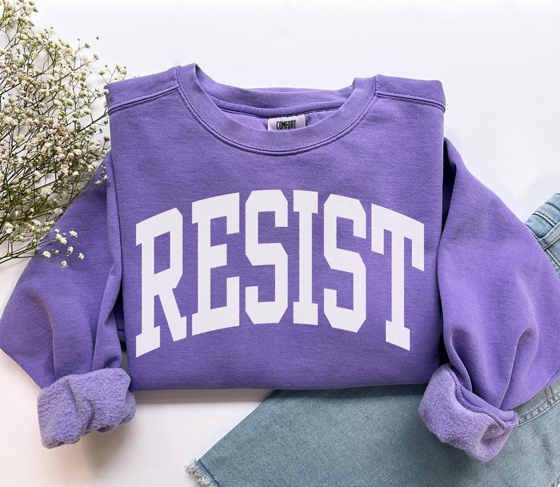 May include: A lavender crewneck sweatshirt with the word "RESIST" in bold white letters. The sweatshirt has a comfortable, casual style, with cuffed sleeves and a relaxed fit. The garment is paired with blue denim jeans.