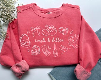 May include: A coral-colored sweatshirt with white graphic illustrations of swans, rings, a ring box, a bow, a cake, a cupid, a champagne bottle, and a love letter. The text "single & bitter" is in the center of the design.
