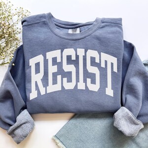 May include: A blue sweatshirt with the word "RESIST" in large, white, block letters across the front. The sweatshirt has a crew neck and ribbed cuffs. The garment is folded on top of a pair of blue jeans.