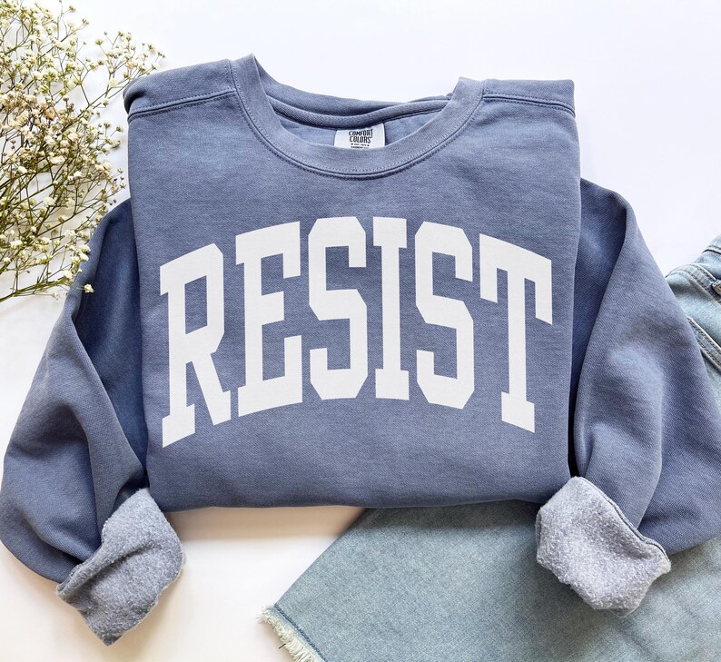 May include: A blue sweatshirt with the word "RESIST" in large, white, block letters across the front. The sweatshirt has a crew neck and ribbed cuffs. The garment is folded on top of a pair of blue jeans.