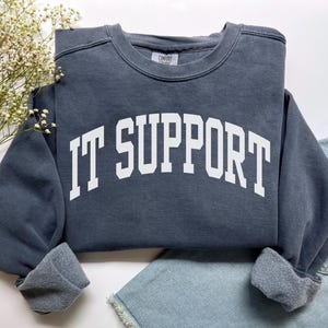 Puede incluir: Una sudadera gris oscuro con las palabras "IT SUPPORT" en letras blancas grandes. La sudadera de cuello redondo tiene puños acanalados. La prenda está doblada sobre una superficie blanca con una ramita de flores blancas y unos shorts vaqueros.