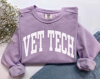 Puede incluir: Una sudadera de color lila con el texto "VET TECH" en letras blancas. La sudadera tiene cuello redondo y mangas largas con puños acanalados. La prenda está doblada sobre un par de vaqueros azules.