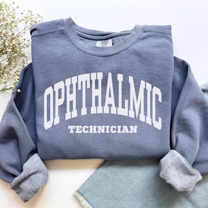 Puede incluir: Una sudadera de cuello redondo en color azul pizarra con el texto blanco OPHTHALMIC TECHNICIAN impreso en una fuente universitaria arqueada y negrita en el pecho. La prenda tiene mangas largas con puños acanalados y forro polar visible.