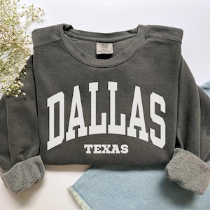 May include: A dark gray sweatshirt with the word "DALLAS" in large white block letters across the chest, and "TEXAS" in smaller white letters below. The sweatshirt has a crew neck and cuffed sleeves.