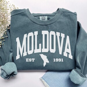 May include: A blue-grey crewneck sweatshirt featuring the white text MOLDOVA in a large arched collegiate font. Below the text, the word EST, a white silhouette map of Moldova, and the year 1991 are printed in white. A Comfort Colors label is visible.