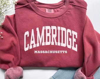 Puede incluir: Una sudadera granate con la palabra "CAMBRIDGE" en letras blancas en bloque arqueadas sobre el pecho, con "MASSACHUSETTS" debajo. La sudadera tiene cuello redondo y puños. Las mangas están enrolladas, revelando un forro rosa más claro.