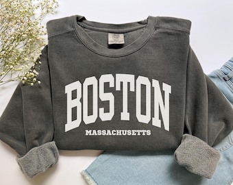Puede incluir: Una sudadera gris oscuro con la palabra "BOSTON" en letras blancas grandes y "MASSACHUSETTS" debajo. La sudadera tiene cuello redondo y mangas con puños. La prenda está doblada sobre unos vaqueros azules.