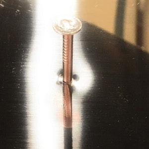 May include: A close-up of a silver nail hammered into a metal surface. The nail is partially visible, with the head and a portion of the shaft showing. The surface is smooth and reflective, creating a mirror-like effect.