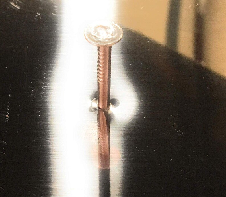 May include: A close-up of a silver nail hammered into a metal surface. The nail is partially visible, with the head and a portion of the shaft showing. The surface is smooth and reflective, creating a mirror-like effect.