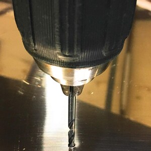 May include: A close-up of a black electric drill with a silver chuck and a black drill bit, drilling into a silver surface.