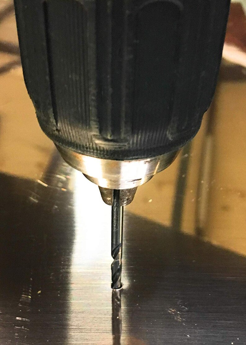 May include: A close-up of a black electric drill with a silver chuck and a black drill bit, drilling into a silver surface.
