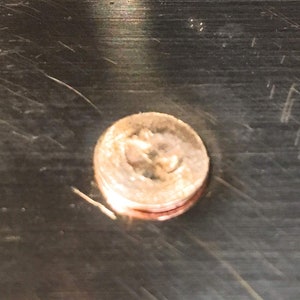 May include: A close-up of a copper-colored coin with a design of a stylized anchor. The coin is on a dark, textured surface.