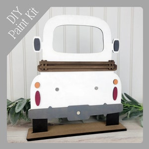May include: A white wooden DIY paint kit shaped like a vintage truck. The truck has a brown bed, gray bumper, and black tires. The text "DIY Paint Kit" is in the upper left corner. The truck is on a brown base.