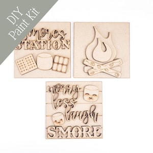 May include: DIY paint kit with three wooden panels. One panel reads "S'mores Station" with marshmallow, biscuit, and chocolate designs. Another panel depicts a campfire. The third panel says "Worry Less Laugh S'more."