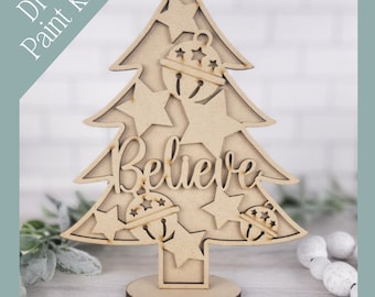 May include: A wooden Christmas tree-shaped DIY paint kit. The tree features star cutouts, bell ornaments, and the word "Believe". The kit includes a base for standing. The text "DIY Paint Kit" is in the upper left corner.
