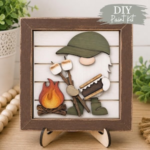 May include: A square wooden sign featuring a layered cutout of a gnome wearing a green hat, holding a s'more and roasting marshmallows over a painted campfire. The artwork is set against a white shiplap background within a dark brown frame.