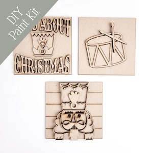 May include: A DIY paint kit featuring three wooden cutouts. One cutout reads "All About Christmas" with a crown design. Another shows a drum with drumsticks, and the third is a nutcracker. The kit is perfect for holiday crafting.