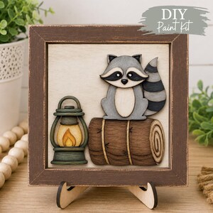 May include: A square wooden framed sign featuring a layered cutout design of a grey and white raccoon sitting on a brown log next to a green lantern with a yellow flame. The sign has a light-coloured background and a dark brown distressed frame.