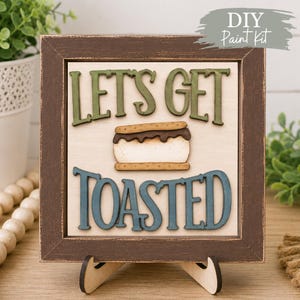 May include: A square wooden sign with a dark brown distressed frame, featuring the text "LET'S GET TOASTED" in raised green and blue letters. A central wooden s'more graphic sits between the lines of text on a light cream background.