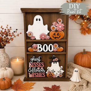 Könnte beinhalten: Ein hölzernes Etagenregal mit Halloween-Motiven. Das oberste Regal zeigt ein weißes Gespenst und einen orangefarbenen Kürbis mit rosa Blumen. Das mittlere Regal zeigt das Wort BOO mit Kürbissen. Das untere Regal enthält ein Gespenst mit Hexenhut und den Text ghost KISSES & flower WISHES.