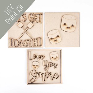 May include: DIY paint kit with three wooden square plaques. One plaque has the text "LET'S GET TOASTED" with crossed marshmallow sticks. Another says "Love You S'more". The plaques feature marshmallow designs.