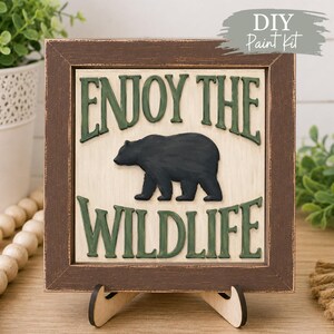 May include: A square wooden sign with a distressed brown frame, featuring a cream-coloured centre panel with raised green lettering that reads ENJOY THE WILDLIFE and a central black bear silhouette. The sign rests on a small wooden stand.