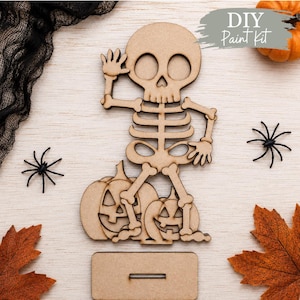 May include: A light brown wooden cutout of a skeleton waving, standing on two jack-o'-lanterns, with a rectangular base. The text "DIY Paint Kit" is visible in the upper right corner. The skeleton has large eye sockets and a rib cage design.