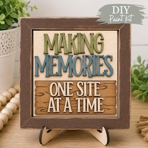 May include: A square wooden sign in a distressed dark brown frame featuring the text "MAKING MEMORIES" in green and blue raised letters, and "ONE SITE AT A TIME" in white letters on a brown wood-grain background, supported by a wooden stand.