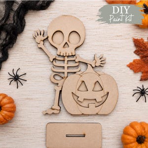 May include: A light brown wooden DIY Paint Kit cutout featuring a cartoon skeleton waving one hand while standing next to a jack-o'-lantern pumpkin. The set includes a rectangular wooden base with a slot for standing the assembled figure upright.