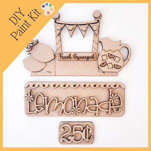 May include: DIY paint kit featuring a wooden lemonade stand design. Includes a sign that says "Lemonade", a pitcher, lemons, and a price tag of 25¢. The kit is unfinished and ready to paint.