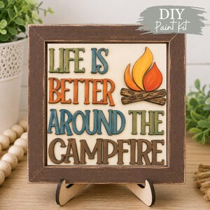 May include: A square wooden sign in a distressed dark brown frame featuring the text "LIFE IS BETTER AROUND THE CAMPFIRE" in raised, multi-coloured letters. A painted graphic of a campfire with orange and yellow flames sits on wooden logs.