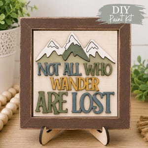 May include: A square wooden sign in a dark brown distressed frame featuring a layered mountain graphic and the text "NOT ALL WHO WANDER ARE LOST" in blue, yellow, and green. The sign is supported by a light-coloured wooden stand.