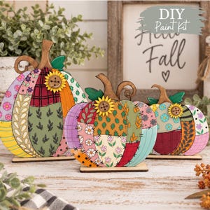 May include: Three wooden pumpkin cutouts featuring patchwork-style patterns in various colors including yellow, purple, red, and green. Each pumpkin is topped with a small wooden sunflower with a button center and stands on a flat base.