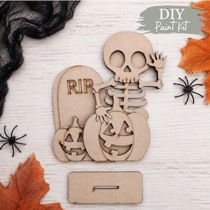 May include: A light brown wooden DIY paint kit featuring a skeleton waving a hand, two jack-o'-lantern pumpkins, and a tombstone with the text RIP. The set includes a rectangular base with a slot for assembly, crafted from flat wood pieces.