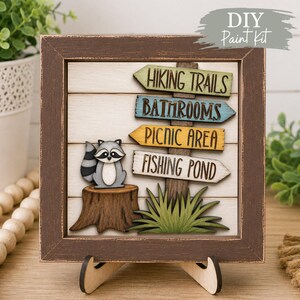 May include: A square wooden sign in a dark brown frame featuring a layered cutout design. It displays a grey and white raccoon sitting on a brown tree stump next to four directional signs labelled HIKING TRAILS, BATHROOMS, PICNIC AREA, and FISHING POND.