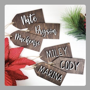 May include: Wooden gift tags with names in white script: Nate, Bryson, Mackenzie, Miley, Cody, and Marina. The tags are brown with a jute string for hanging. A red poinsettia and evergreen sprig with red berries are in the background.