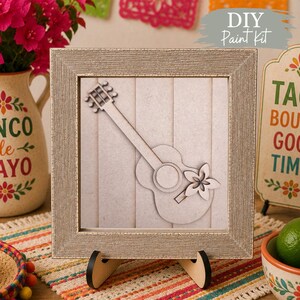 Peut inclure: Un cadre carré en argent texturé contenant une découpe en bois clair représentant une guitare avec un détail floral. La guitare est superposée sur un fond à planches verticales. Le cadre repose sur un support noir et le texte DIY Paint Kit est visible.