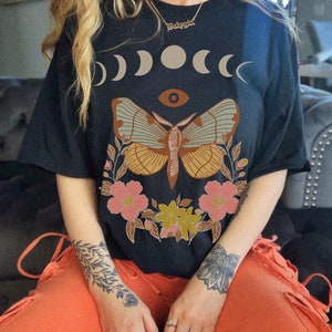 以下が含まれることがあります： 三日月と目のデザインに、花柄のアクセントが施された蛾のグラフィックプリントが施された、黒色のオーバーサイズTシャツ。ゴールドチェーンネックレスに「Babygirl」の文字が見える。