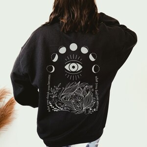 Op de afbeelding: Zwart sweatshirt met een witte afbeelding op de rug. De afbeelding bevat een oog, maanfasen en botanische elementen. De tekst luidt: "wat je denkt word je, wat je voelt trek je aan."