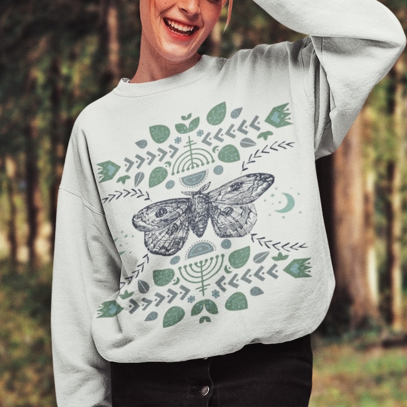 May include: A white sweatshirt with a green and blue geometric pattern featuring a moth in the center. The pattern includes leaves, arrows, and other abstract shapes.
