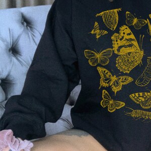 May include: A black long-sleeved sweatshirt with a gold print of various butterflies and insects. The sweatshirt is worn by a person sitting on a gray couch.