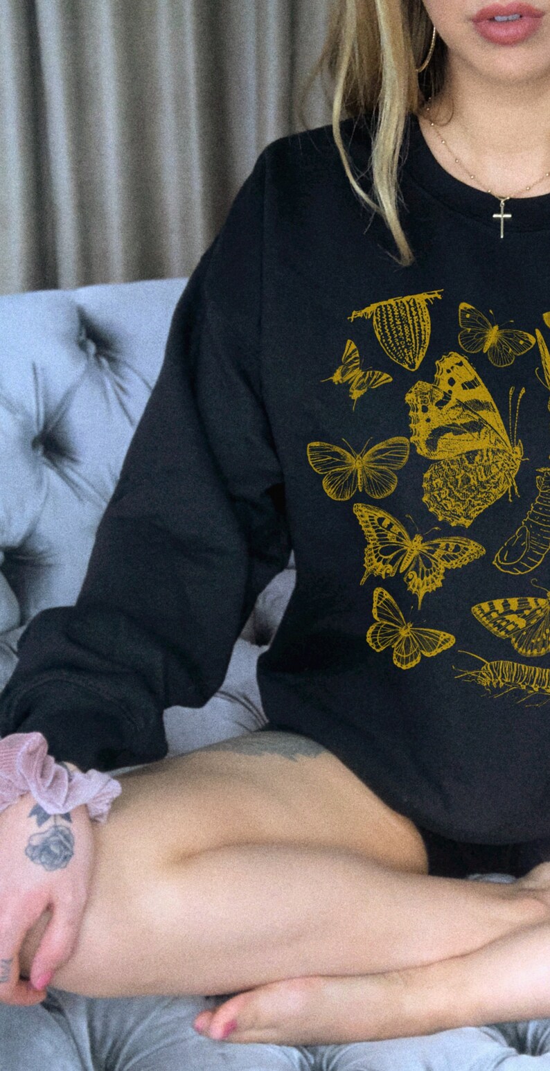 May include: A black long-sleeved sweatshirt with a gold print of various butterflies and insects. The sweatshirt is worn by a person sitting on a gray couch.
