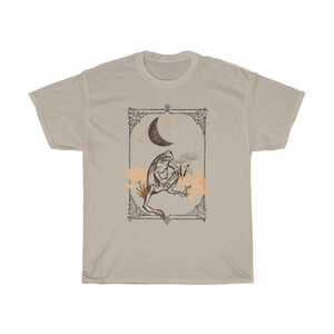 K&ouml;nnte beinhalten: Ein beiges T-Shirt mit einer Vintage-Illustration einer unter einem Halbmond sitzenden Kr&ouml;te. Die Illustration ist mit einem dekorativen Rahmen versehen und zeigt kleine Blumen und Sterne.