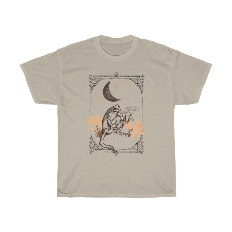 K&ouml;nnte beinhalten: Ein beiges T-Shirt mit einer Vintage-Illustration einer unter einem Halbmond sitzenden Kr&ouml;te. Die Illustration ist mit einem dekorativen Rahmen versehen und zeigt kleine Blumen und Sterne.