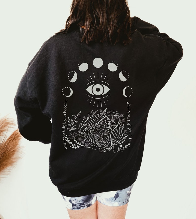 Op de afbeelding: Zwart sweatshirt met een witte afbeelding op de rug. De afbeelding bevat een oog, maanfasen en botanische elementen. De tekst luidt: "wat je denkt word je, wat je voelt trek je aan."