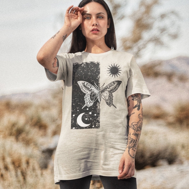 May include: A light gray t-shirt with a black and white graphic print of a butterfly, a crescent moon, and a sun. The graphic is on a distressed background.